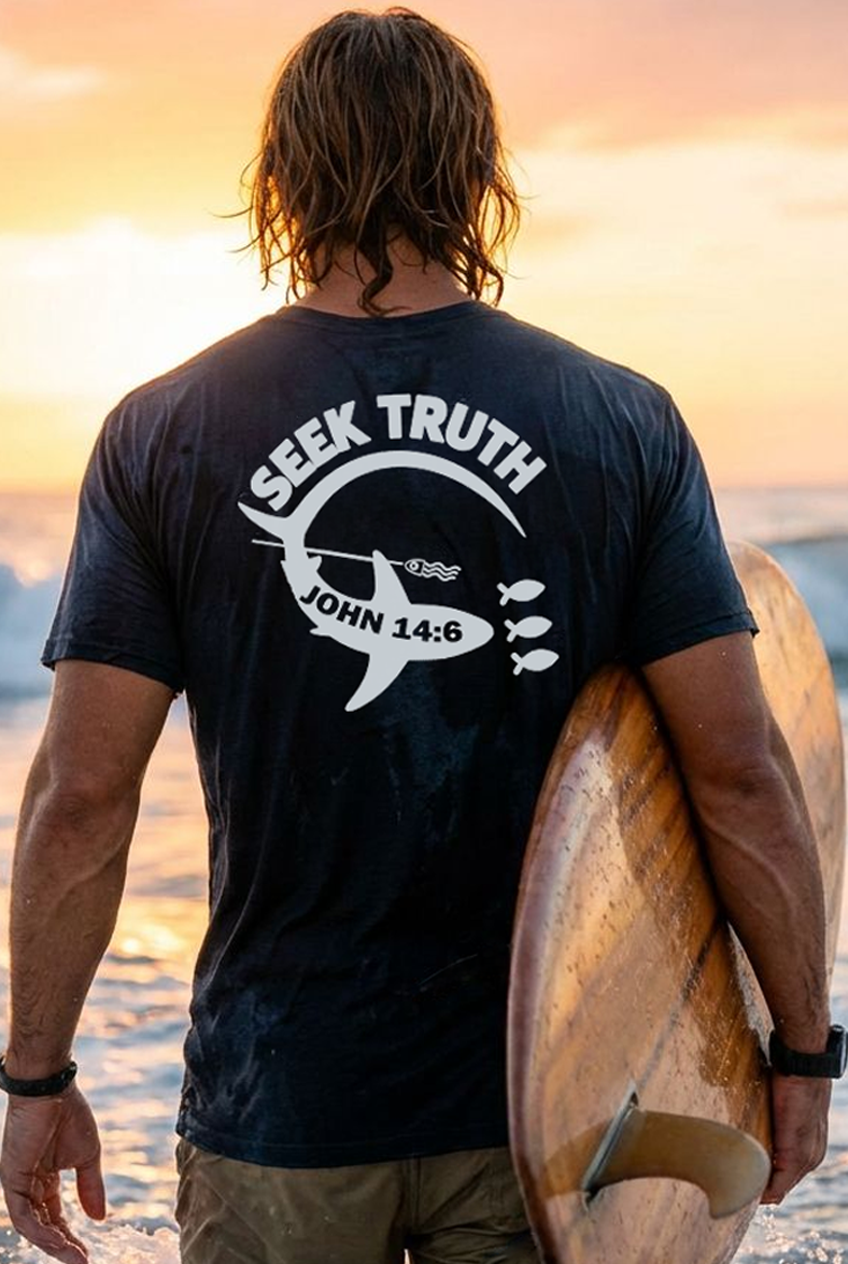 Person wearing a black t-shirt with a white shark, 'Seek Truth' and 'John 14:6' text and holding a surfboard at sunset.