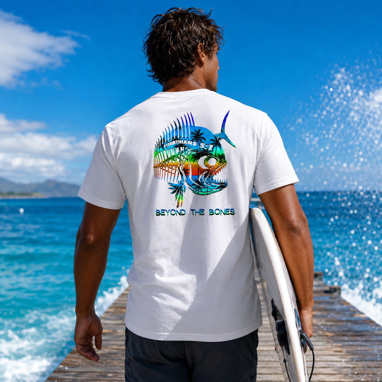 Man wearing a white t-shirt with a colorful fish design and 'Beyond the Bones' text, holding a surfboard on a wooden pier with ocean view.