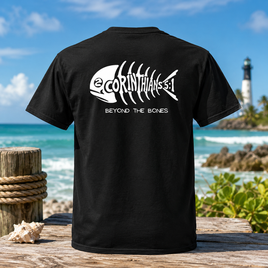 A black short-sleeve T-shirt shown from the back, featuring a white fish skeleton graphic with the text '2CORINTHIANS 5:1' and 'BEYOND THE BONES.' The shirt is photographed outdoors on a wooden surface with a beach, ocean, and lighthouse in the background.