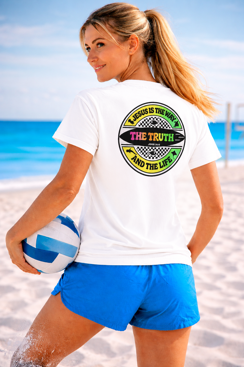 Woman on a beach holding a volleyball, wearing a white t-shirt with a colorful graphic and text.