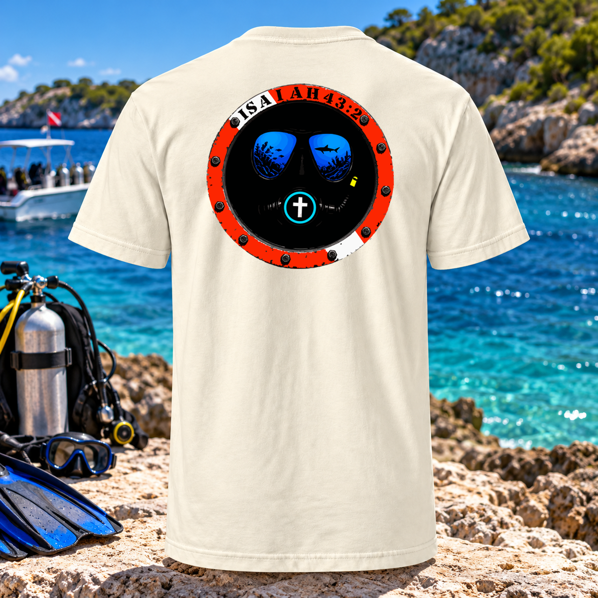 A cream-colored t-shirt displayed on a rocky beach with a tropical ocean background. The back of the shirt features a circular graphic resembling a submarine or diving helmet with the text 'ISALAH 44:23' in white on a red ring, blue water reflections inside, and a white cross in the center.