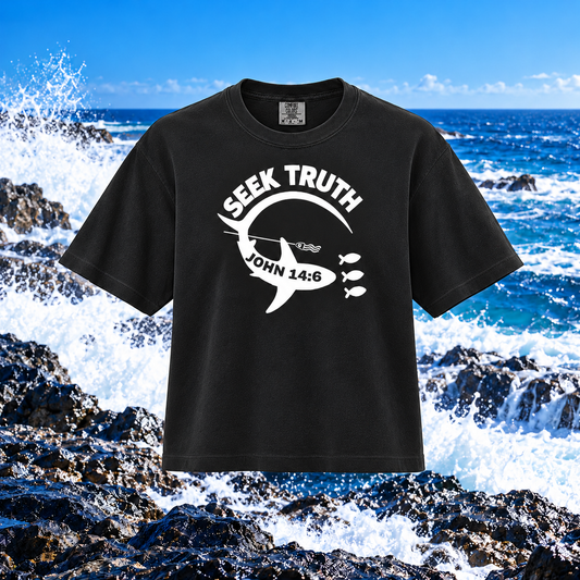 A black short-sleeve graphic T-shirt with a white shark design and the text 'SEEK TRUTH JOHN 14:6' on the front, shown against a rocky ocean background with waves crashing.