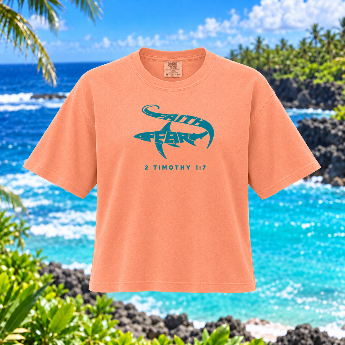 A peach-colored short-sleeve t-shirt with a teal graphic of a shark containing the words 'GOD FEARS' and the text '2 TIMOTHY 1:7' printed below it. The shirt is displayed against a tropical beach background with blue ocean water, rocks, and palm trees.