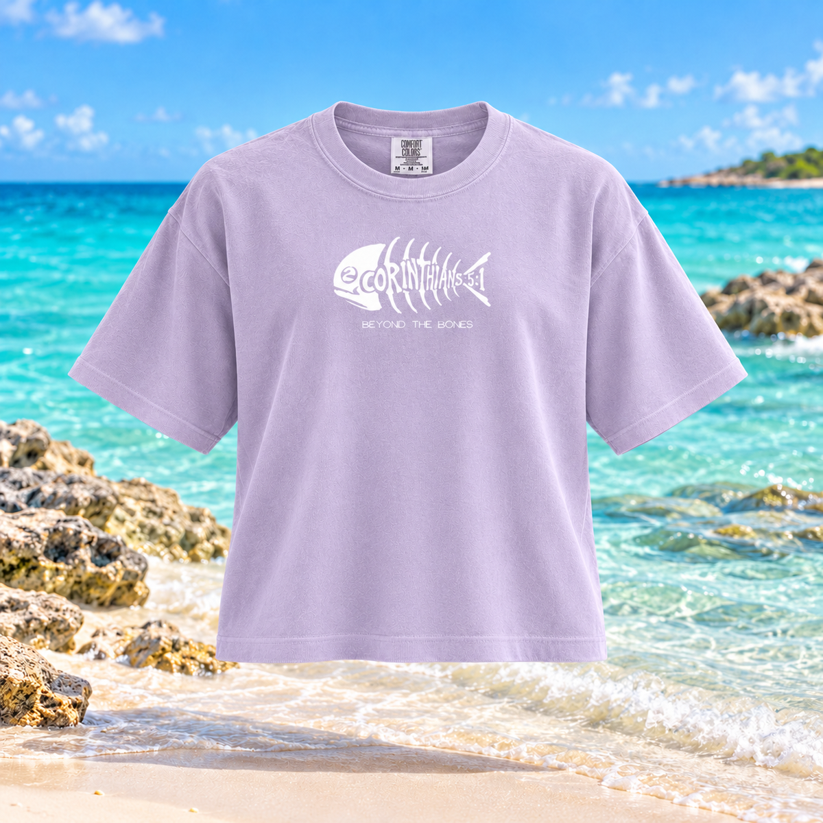 A lavender crewneck t-shirt with a white fish skeleton graphic and the text 'CORINTHANS BEYOND THE BONES' on the chest, displayed against a tropical beach background with turquoise water and rocks.