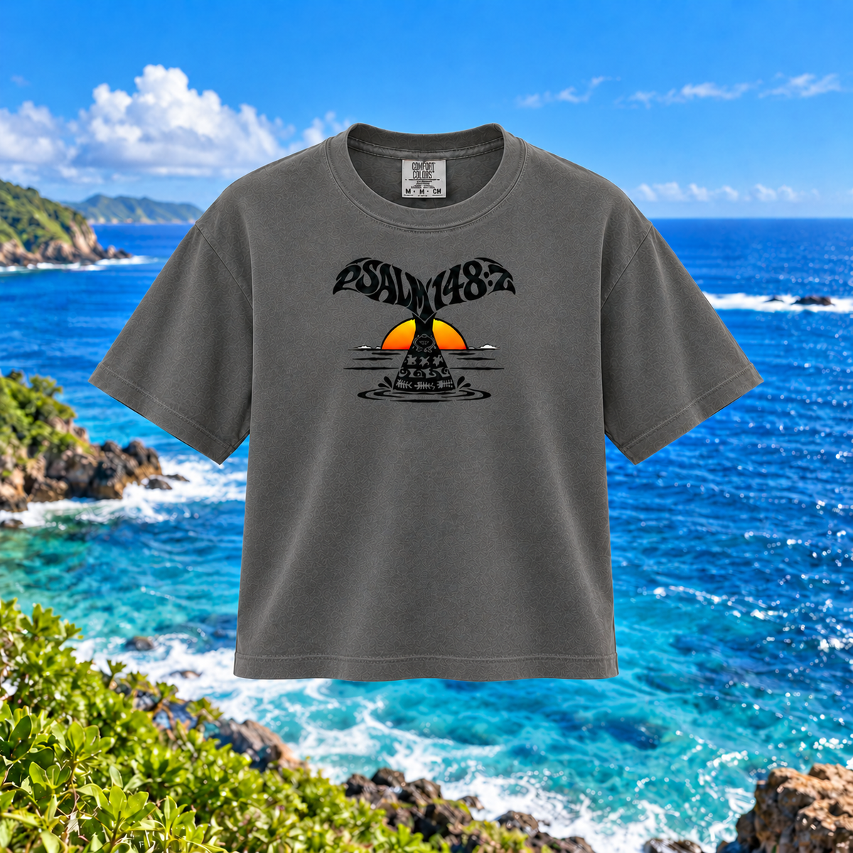 A dark gray graphic t-shirt featuring a whale tail design with the text 'ESALY 148' and a yellow-orange sun over water, shown against a scenic ocean background.