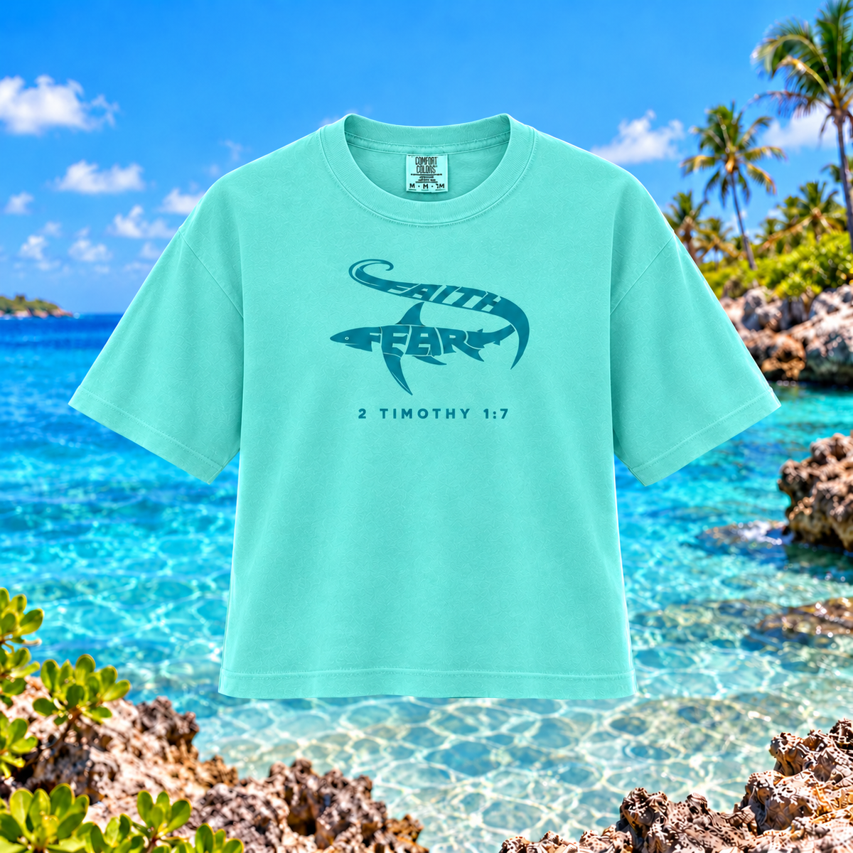 A mint green short-sleeve t-shirt with a dark teal graphic of a shark and the words 'SEATH FEAR' and '2 TIMOTHY 1:7' printed on the chest, displayed against a tropical beach background.
