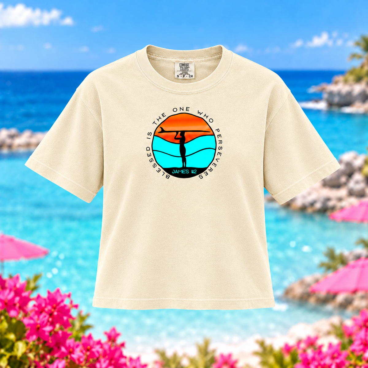 A cream-colored short-sleeve t-shirt with a circular graphic on the chest, displayed against a blurred beach background with turquoise water, pink umbrellas, and flowers.