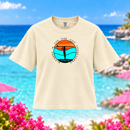 A cream-colored short-sleeve t-shirt with a circular graphic on the chest, displayed against a blurred beach background with turquoise water, pink umbrellas, and flowers.