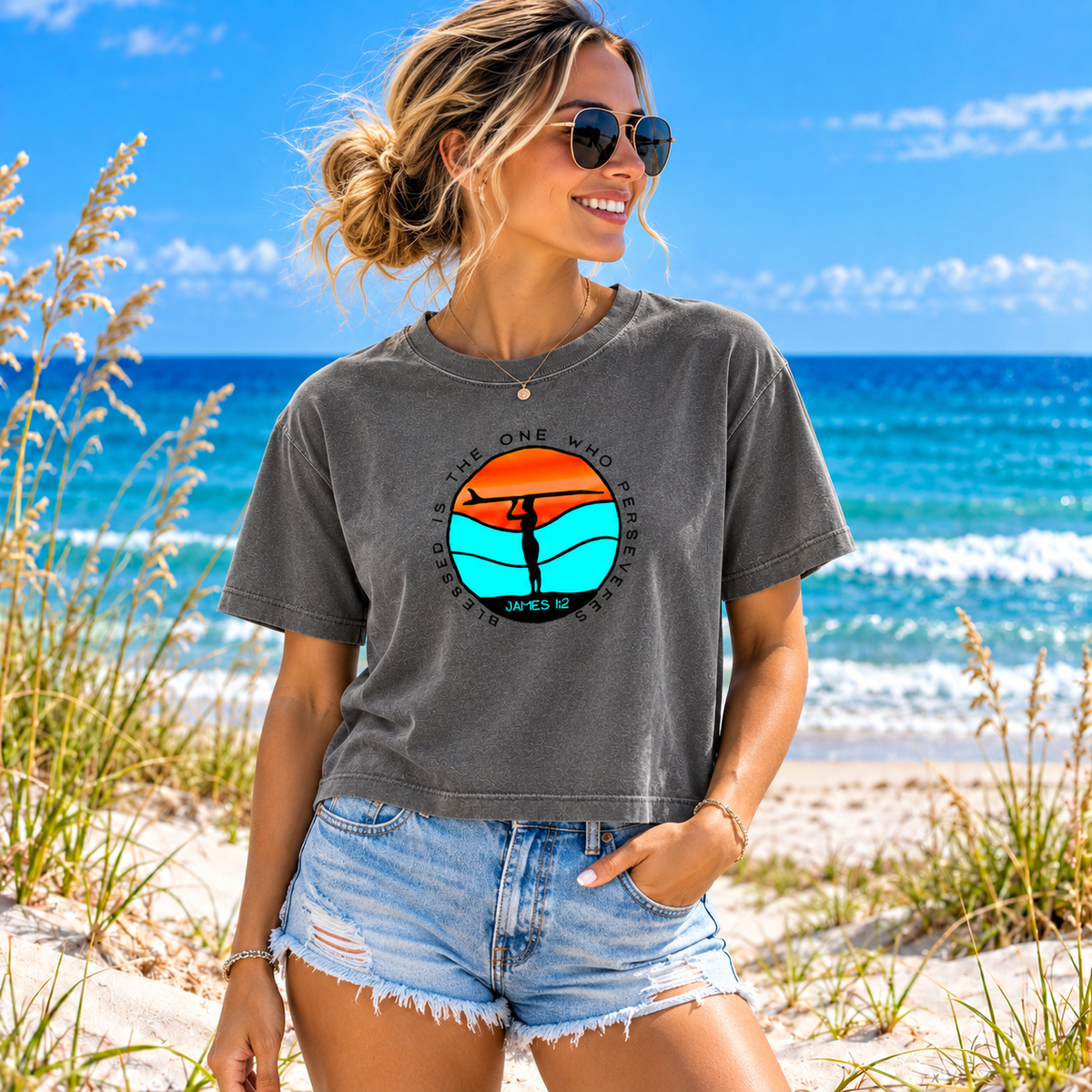 A woman wearing a dark gray graphic t-shirt and denim shorts stands on a sandy beach with the ocean and blue sky in the background. The t-shirt features a circular graphic with a sunset and a silhouette of a person, with the text 'IF YOU ARE ONE WHO DESPIRES, JUDGE ONESELF AS DESPIRED' and 'JUNE 12'.