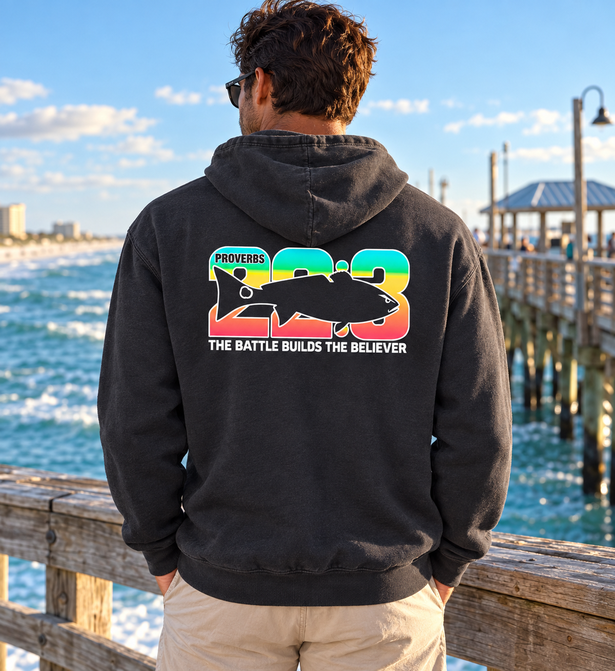 A man viewed from behind wearing a black hoodie with a large back graphic featuring the number 23 in rainbow colors, a fish silhouette, and the text 'PROVERBS 23 THE BATTLE BUILDS THE BELIEVER'. He is standing on a wooden boardwalk with the ocean and a pier in the background.