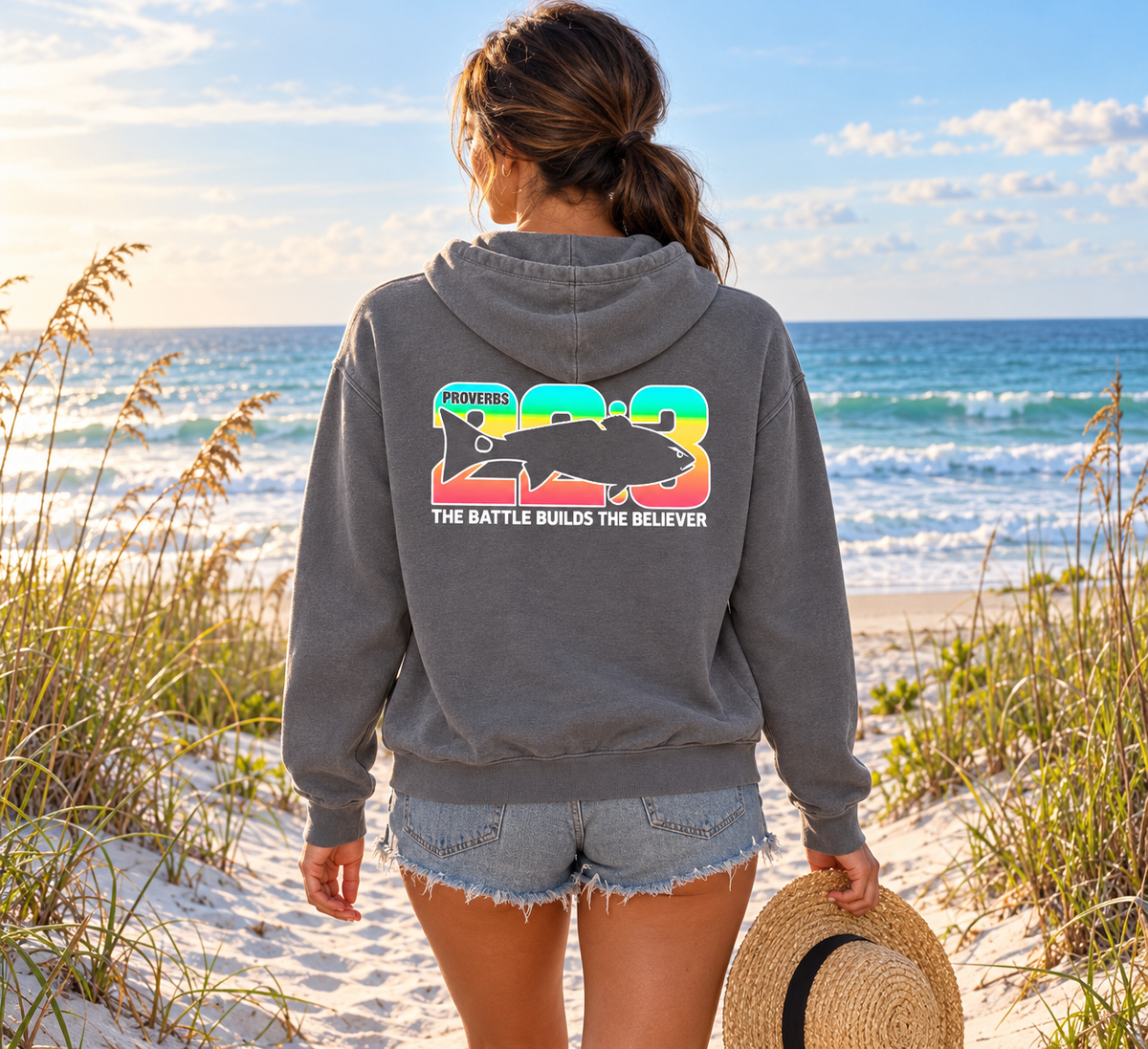 A woman stands on a sandy beach facing the ocean, wearing a heather gray hoodie with a colorful graphic on the back that reads 'FISHING WITH THE BATTLE BUILDS THE BELIEVER' and '1993'. She is also wearing denim shorts and holding a straw hat.