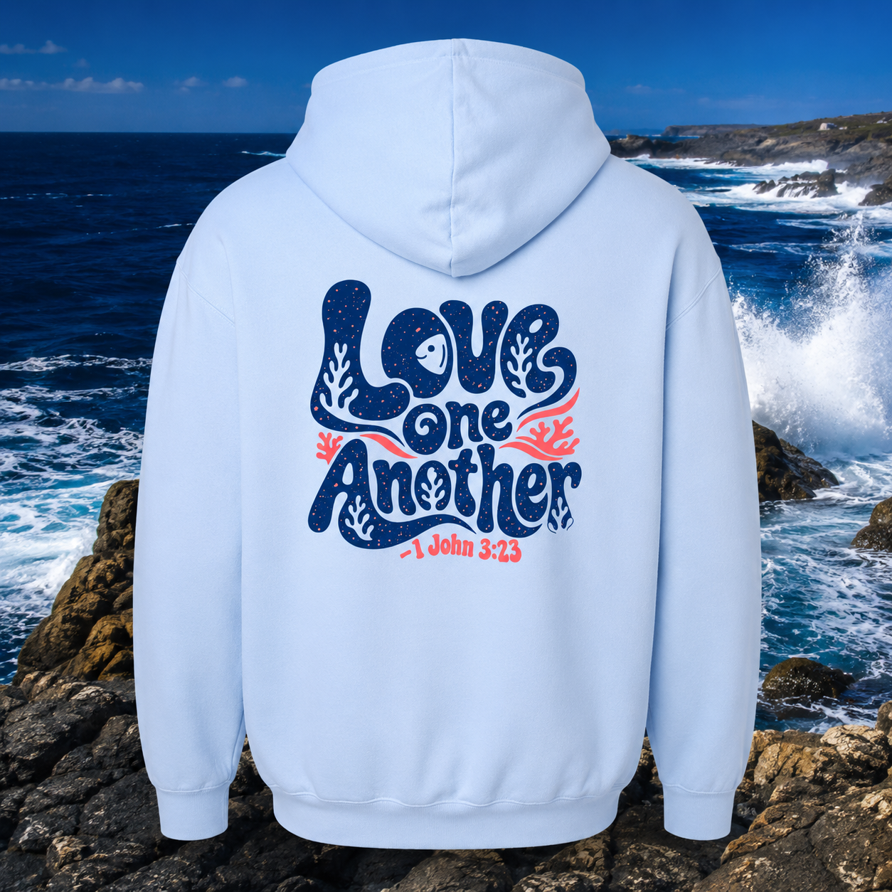 White hoodie with 'Love One Another' text on the back, worn by a person standing on rocks with ocean waves in the background.