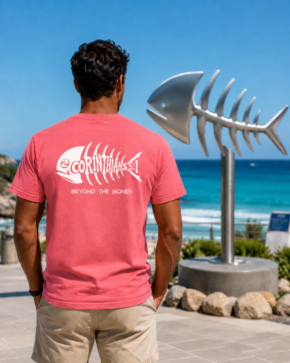 A man viewed from behind wearing a coral pink t-shirt with a white fish skeleton graphic and the text '2CORINTHIANS-5:1 BEYOND THE BONES' on the back. He is standing outdoors on a sunny day with the ocean, beach, and a large metallic fish skeleton sculpture in the background.