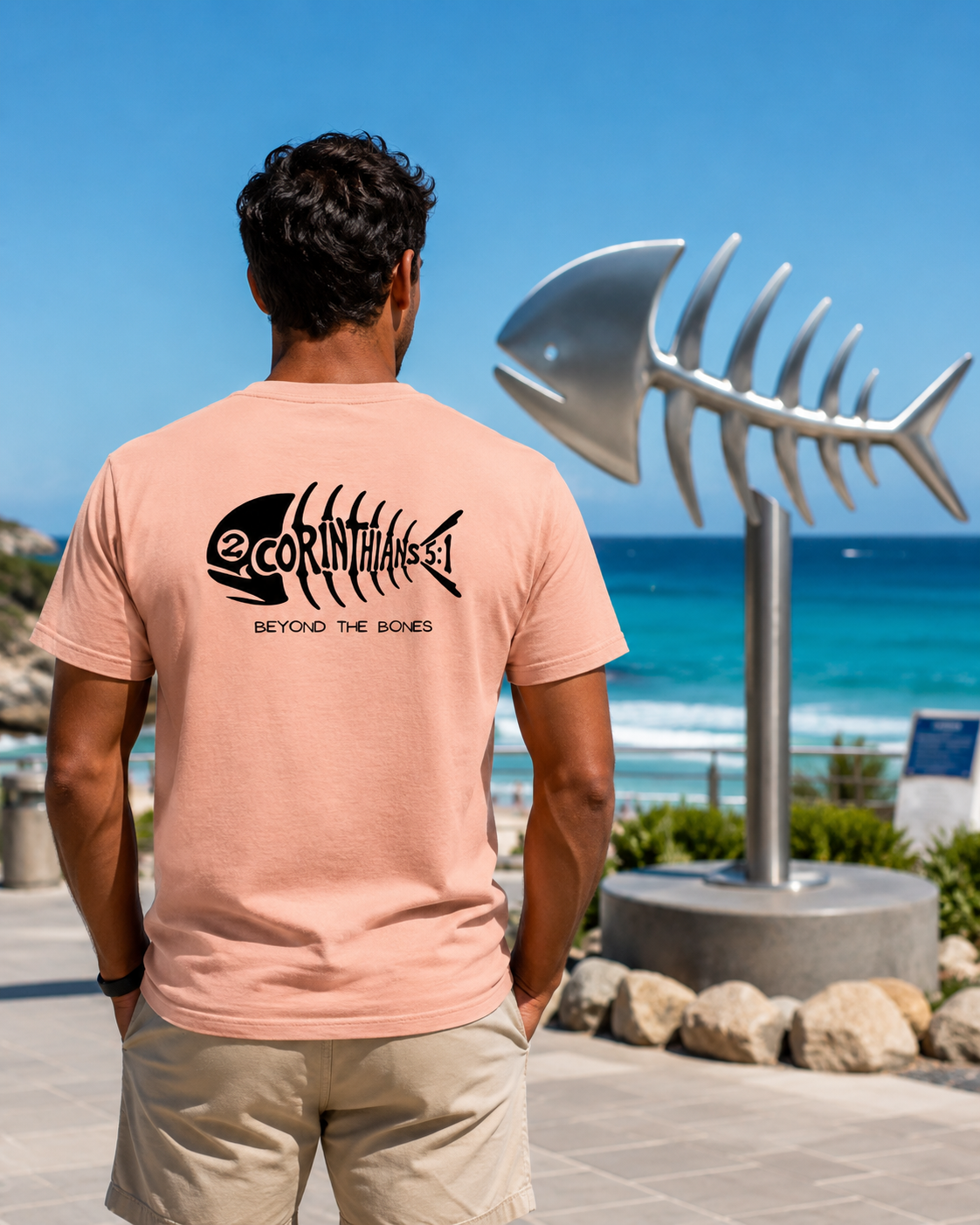 A man viewed from behind wearing a peach-pink t-shirt with a black fish skeleton graphic and the text '2Corinthian5:1' BEYOND THE BONES'. He is standing outdoors on a paved walkway next to a large metallic fish skeleton sculpture, with a blue ocean and sky in the background.