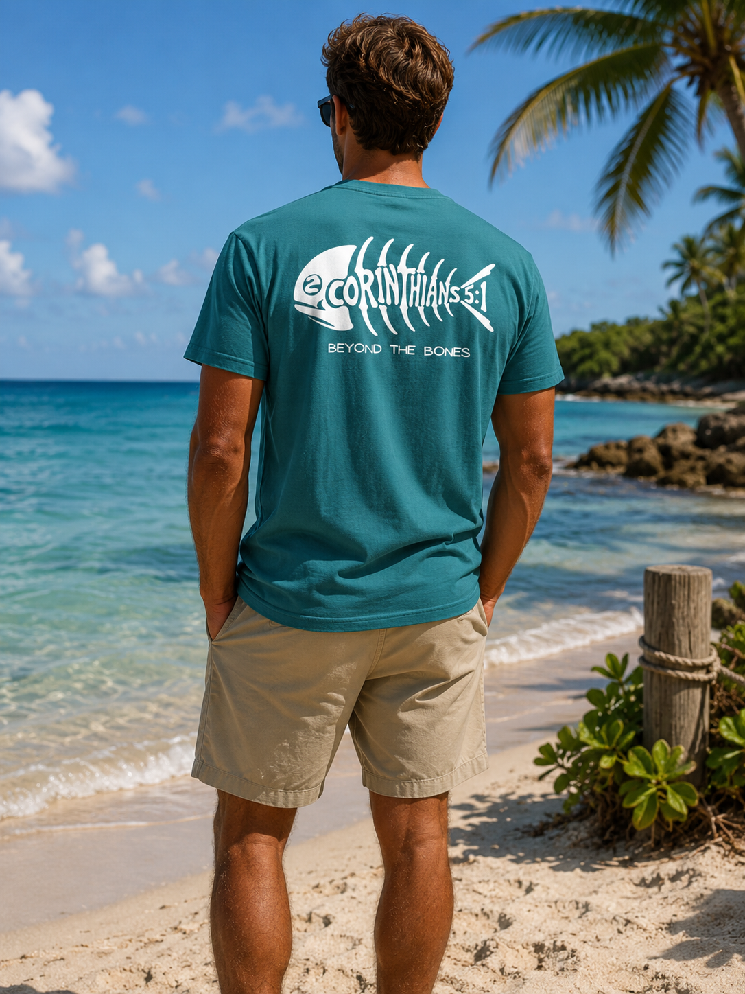A man standing on a sandy beach with the ocean and palm trees in the background, wearing a teal t-shirt with a white fish skeleton graphic and the text '2 CORINTHIANS-5:1' and 'BEYOND THE BONES' on the back, paired with khaki shorts.
