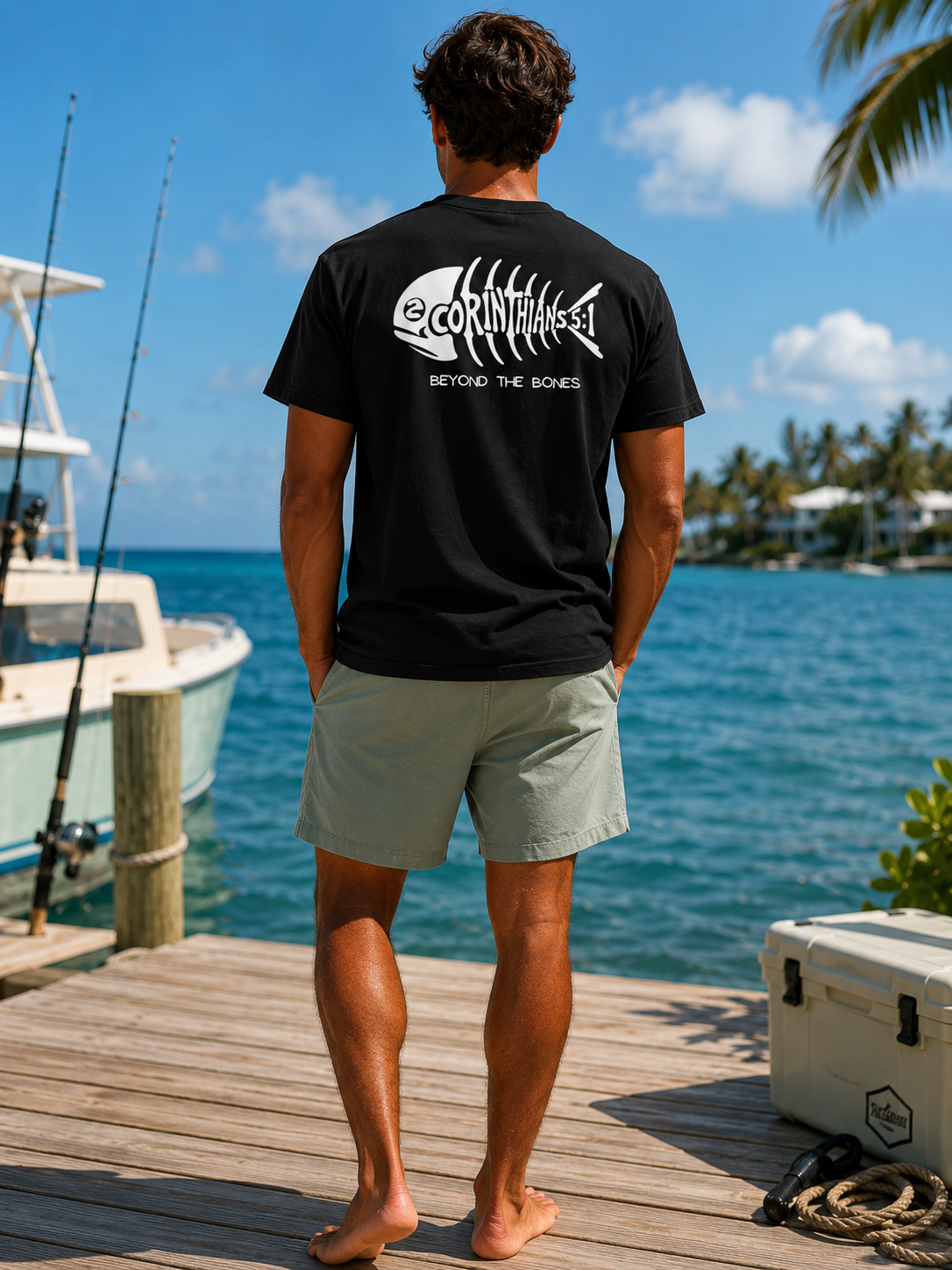 A man stands on a wooden dock by the water wearing a black t-shirt with a white fish skeleton graphic and the text '2CORINTHIANS5:1' and 'BEYOND THE BONES'.