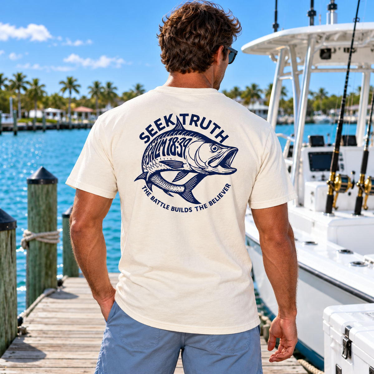 A man standing on a wooden dock by a boat, viewed from behind, wearing a cream-colored t-shirt with a large navy blue fish graphic and the text 'SEEK TRUTH FISHING' and 'THE BATTLE BUILDS THE BELIEVERS' on the back.