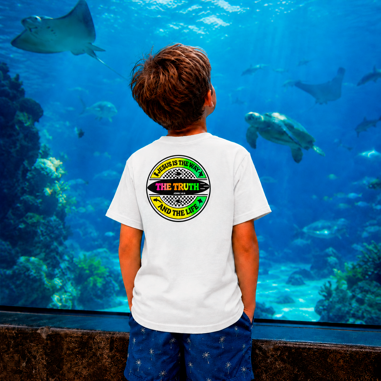 Child wearing a white t-shirt with a colorful design in front of an aquarium with fish and coral.